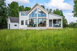 A two-story house with a large glass front and a porch with chairs. - The Birches on Marbles Way | Snowmobile Access | Waterfront Access on Rangeley Lake (Rangeley)