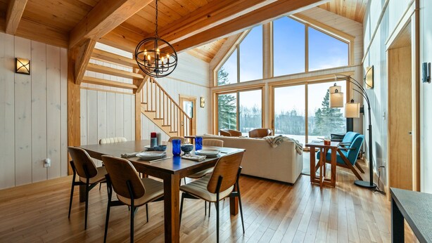 A spacious dining area with a wooden table and chairs, a staircase leading to an upper level, and large windows offering a view of the outdoors. - The Birches on Marbles Way | Snowmobile Access | Waterfront Access on Rangeley Lake (Rangeley)