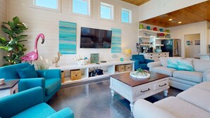 Ein geräumiges Wohnzimmer mit Holzbalkendecke, einem großen Flachbildfernseher, türkisfarbenen Sofas und einer Küchenzeile mit weißen Schränken und einem Kühlschrank.