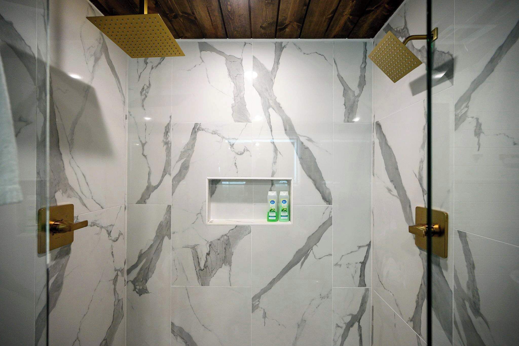 Ein Badezimmer mit Marmorfliesen, einer Dusche inklusive goldfarbener Duschbrause und einem Seifenspender an der Wand.