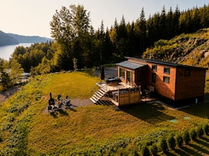 A cabin with a deck and outdoor seating, surrounded by a forest and a lake.