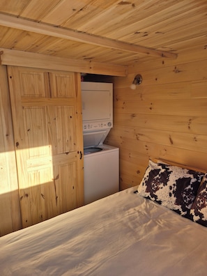 Interior de una cabaña de madera con una cama, una lavadora y una puerta de madera.