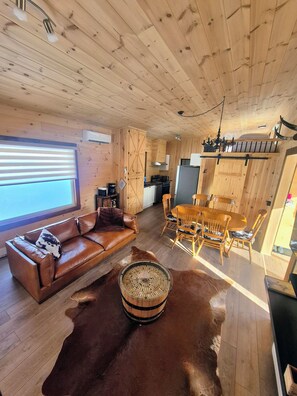 A wooden cabin interior with a leather sofa, dining table, chairs, and a fur rug.