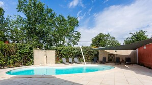 A swimming pool with tiled flooring and lounge chairs, surrounded by greenery and a building with a sloped roof. - The red gîte of the Gîtes de Bel-Air in Luberon (TAILLADES)