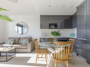 A modern living room with a glass dining table, wooden chairs, a sofa with cushions, a round mirror, and a kitchen with dark cabinets and a refrigerator.