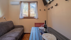 Een kamer met een bank, een eettafel met een fles, een bord en bestek. Er is een raam met uitzicht op bomen en een berg.