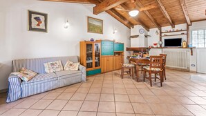 A cozy living room with a striped sofa, a dining table with chairs, a wooden cabinet, and a kitchen area with a stove and a clock. - Casa Rolè, Lake Maggiore Patio House w/ BBQ, Dormelleto, Italy (Dormelleto)