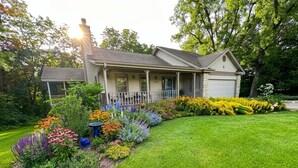 A two-story house with a front porch, surrounded by a well-maintained lawn and colorful flower beds. - Spacious House with King beds, Fire places, Game Room and Dog friendly. (Galena)