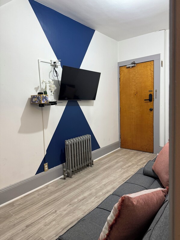 Una habitación con un televisor montado, un radiador, una puerta de madera y un sofá con cojines.