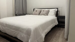 Een net opgemaakt bed met een witte dekbedovertrek en kussens, een nachtkastje met een lamp, en uitzicht op zee door ronde ramen.