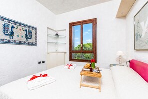 A hotel room with a bed, a small table with breakfast items, a window with a view of greenery, and a decorative wall hanging. - Il Vecchio Faro 66, Porto San Paolo, Italy (Porto San Paolo)