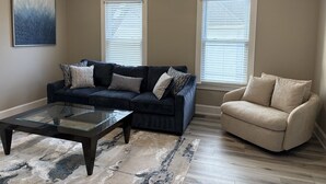 A living room with a dark sofa, beige armchair, glass coffee table, patterned rug, and two windows with blinds. - Spacious Luxury Apartment in Restored Asbury Mansion (Asbury Park)