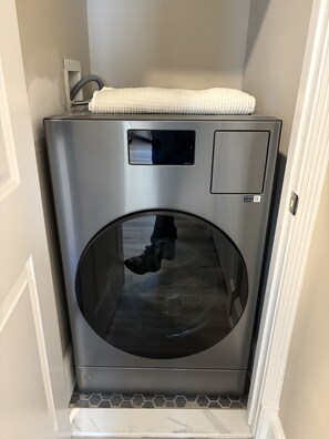 A compact, stainless steel washing machine in a narrow hallway with a tiled floor.