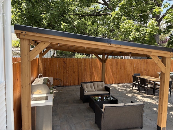 A covered patio area with outdoor furniture, a grill, and a wooden fence.