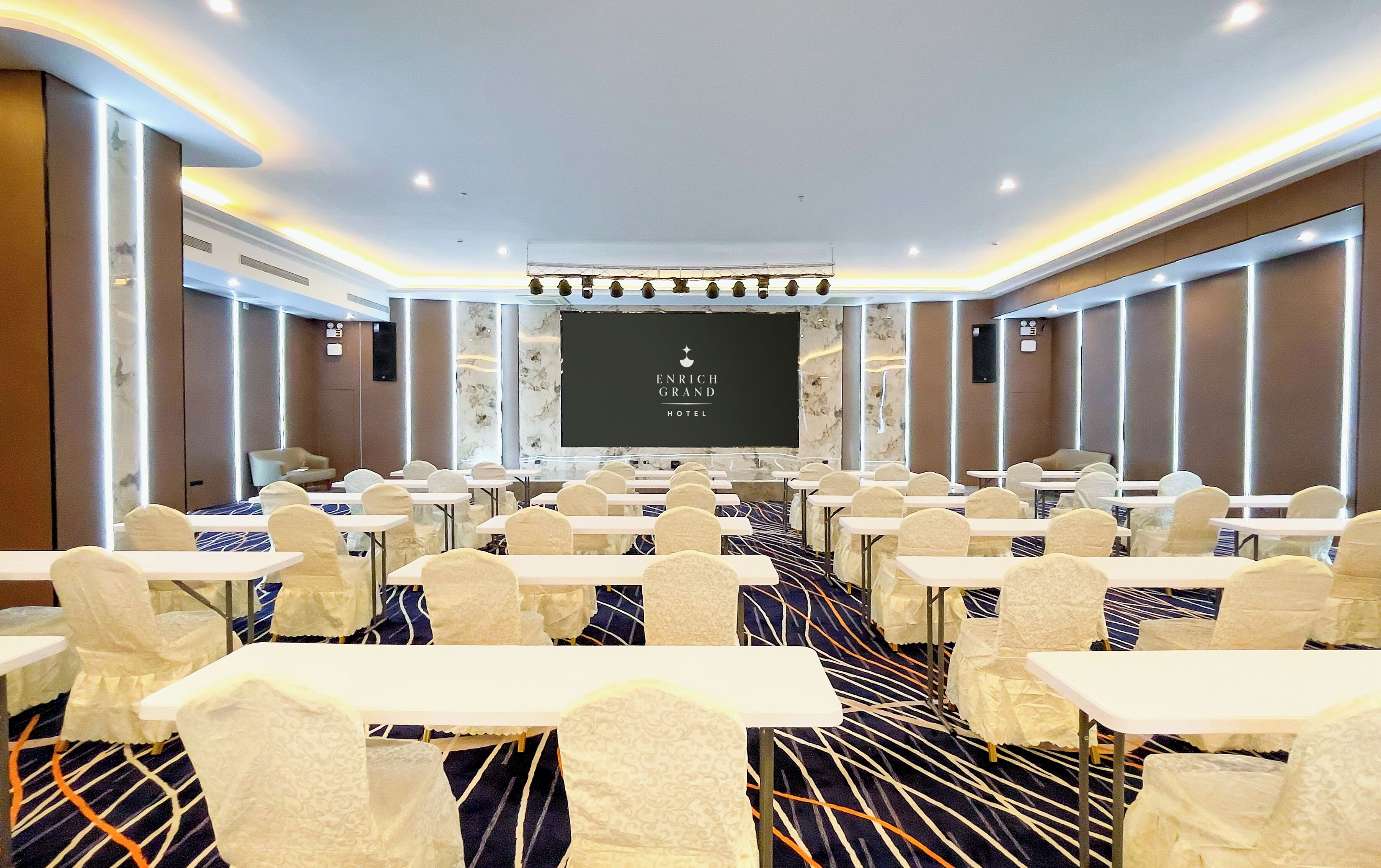 Un salón de conferencias con mesas y sillas, una pantalla grande y el logotipo del hotel Enrich Grand.