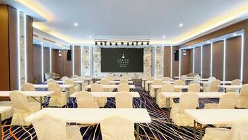 Un salón de conferencias con mesas y sillas, una pantalla grande y el logotipo del hotel Enrich Grand.