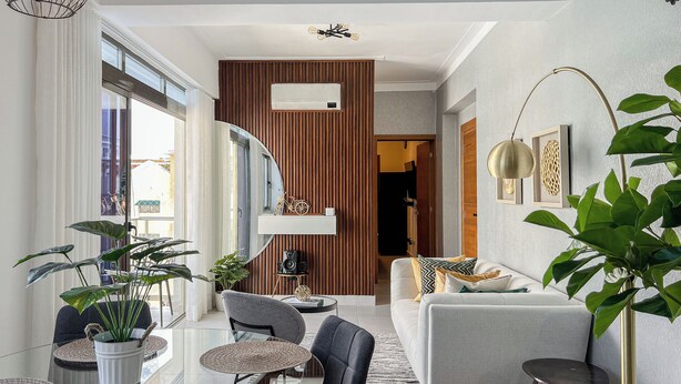 Un salon moderne comprenant un canapé, des fauteuils, une table basse et un coin repas avec une table et des chaises en verre. On y trouve également des plantes, un miroir et une étagère murale agrémentée d’objets décoratifs.