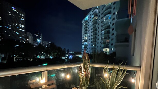 A balcony with a wooden table and chairs, potted plants, and a view of a cityscape at night. - Modern & cozy apartment beachfront (Sunny Isles Beach)