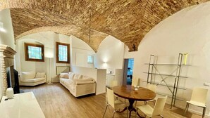 A spacious living area with a high wooden ceiling, a round dining table with chairs, a white sofa, and a small kitchen area with a radiator. - Le Volte di via Portica (Assisi)
