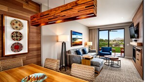 Ein modernes Hotelzimmer mit Holzboden, einer Essecke mit Holztisch, einer Couch, einem Fernseher und einem Balkon mit Bergblick.