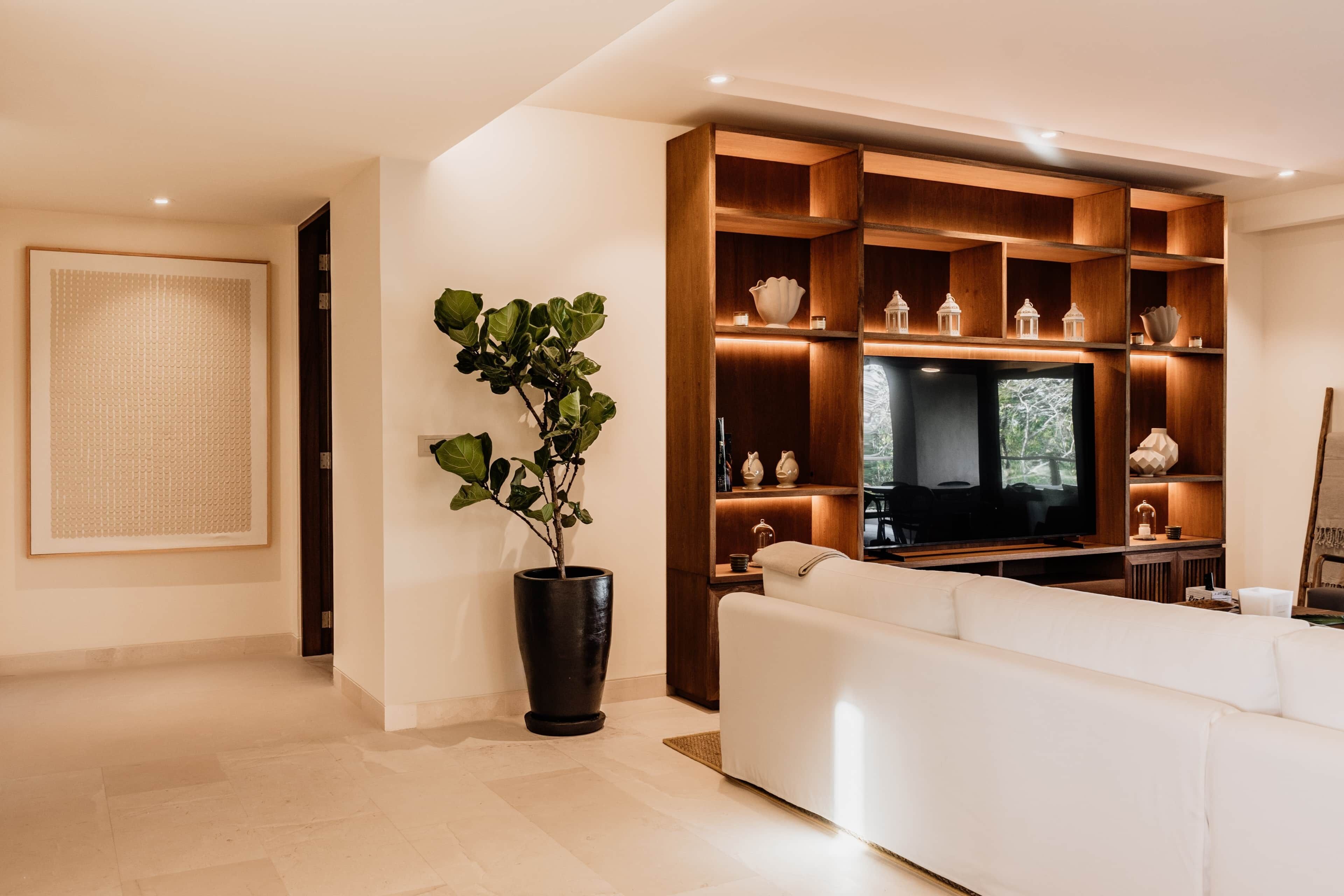 Un salon moderne doté d’un grand téléviseur à écran plat, d’un canapé blanc, d’une étagère en bois agrémentée d’objets décoratifs et d’une plante en pot.
