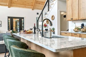 A modern kitchen with a marble countertop, green upholstered bar stools, wooden cabinets, and a staircase with a railing.