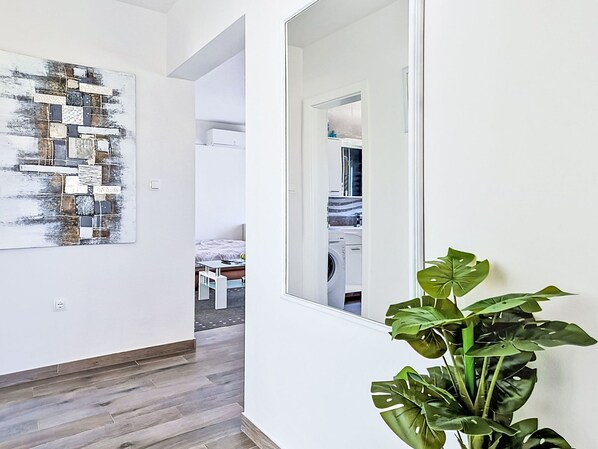 Un couloir avec un plancher en bois, une grande peinture abstraite, une plante en pot et une vue sur une chambre à coucher avec un lit et un lave-linge.