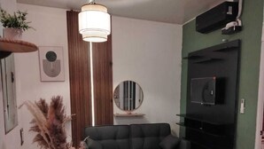 Una sala de estar moderna con una mesa de comedor de cristal, un sofá negro, un televisor montado en la pared y un jarrón decorativo con plantas secas.