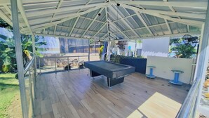 Un espacio al aire libre techado con mesa de billar, asientos y una pérgola.