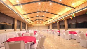 A large hall with rows of tables and chairs, a high wooden ceiling with lighting, and large windows. - Hotel Sparrow (Dehradun)