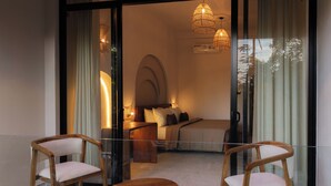 A modern bedroom with a bed, bedside tables, and a glass partition. There are two wooden chairs and a round wooden table in the foreground. - The Benison (Ahangama)