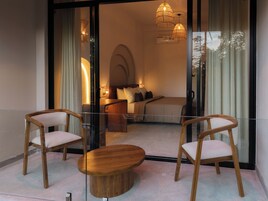 A modern bedroom with a bed, bedside tables, and a glass partition. There are two wooden chairs and a round wooden table in the foreground.