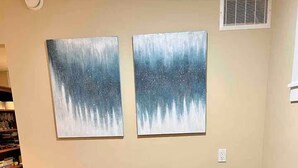 A dining area with a wooden table set for four, two wall-mounted abstract paintings, and a bookshelf in the background. - Beautiful 1 bedroom suite close to UBC (Vancouver)