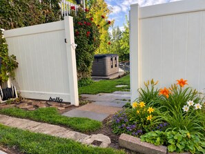 A garden pathway leading to a white gate with a 'hello' sign, surrounded by various flowers and a small shed. - Art City Escape - Luxury Swim Spa & Private Entertainment Retreat (Springville)