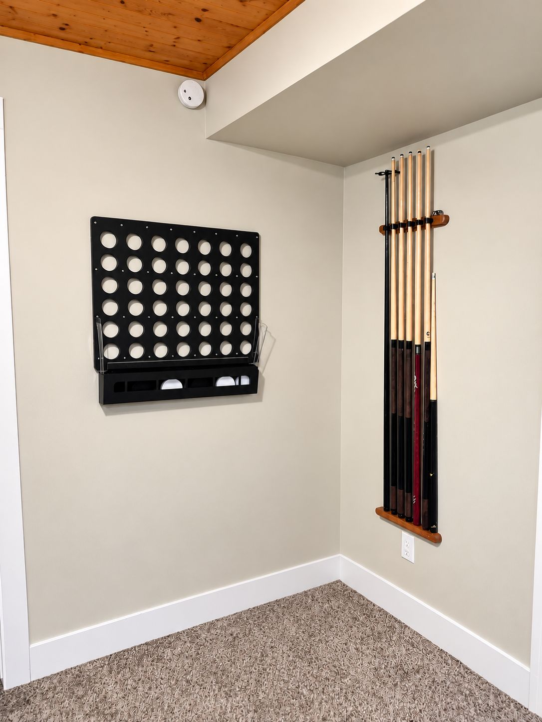 Une pièce avec un tapis beige, un présentoir noir fixé au mur, percé de trous circulaires, et un présentoir vertical pour les queues de billard.