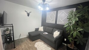 A living room with a sectional sofa, a ceiling fan, decorative wall panels, a mounted deer head, a potted plant, and a television on a stand. - Located in Basement of Twin Home. Relax & Play with Swim Spa & Game Room for 4 (Springville)