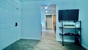A hallway with a wooden floor, a door on the left, a TV on the right, and a plant shelf below it. - Located in Basement of Twin Home. Relax & Play with Swim Spa & Game Room for 4 (Springville)