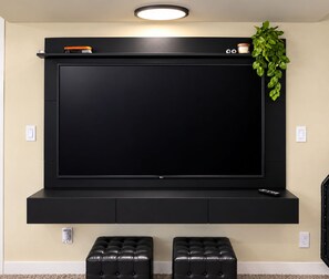 A black entertainment unit with a flat-screen TV, two black ottomans, a small shelf with decorative items, and a green plant.