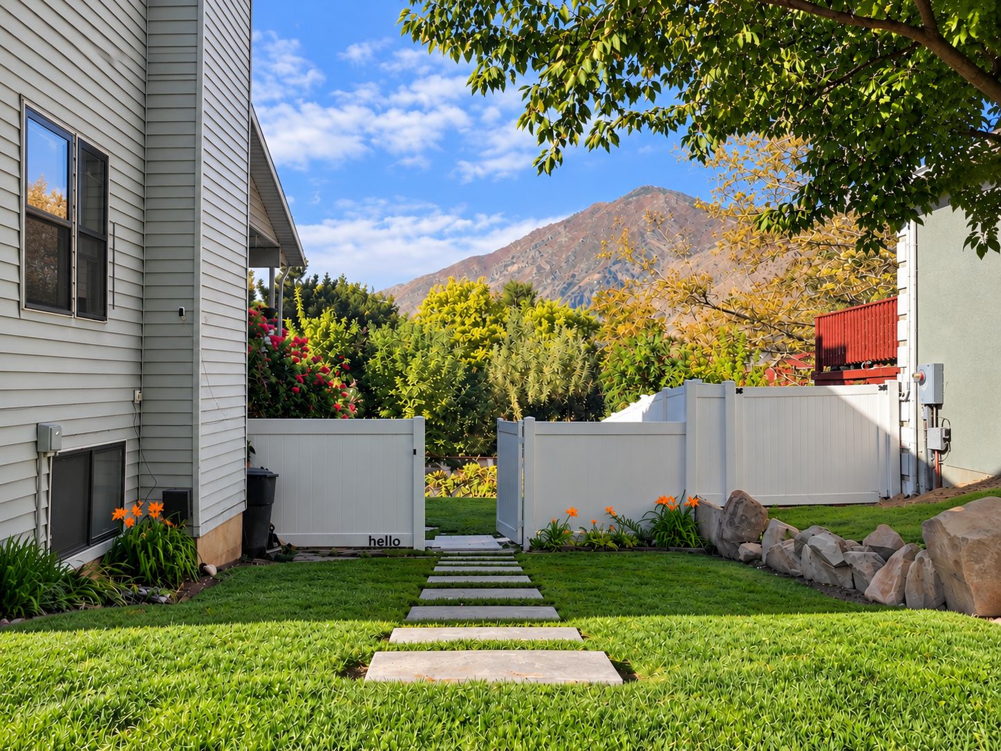 Un jardin bien entretenu, avec un chemin de pierres menant à une porte, entouré d’une végétation luxuriante et dominé par une chaîne de montagnes en arrière-plan.