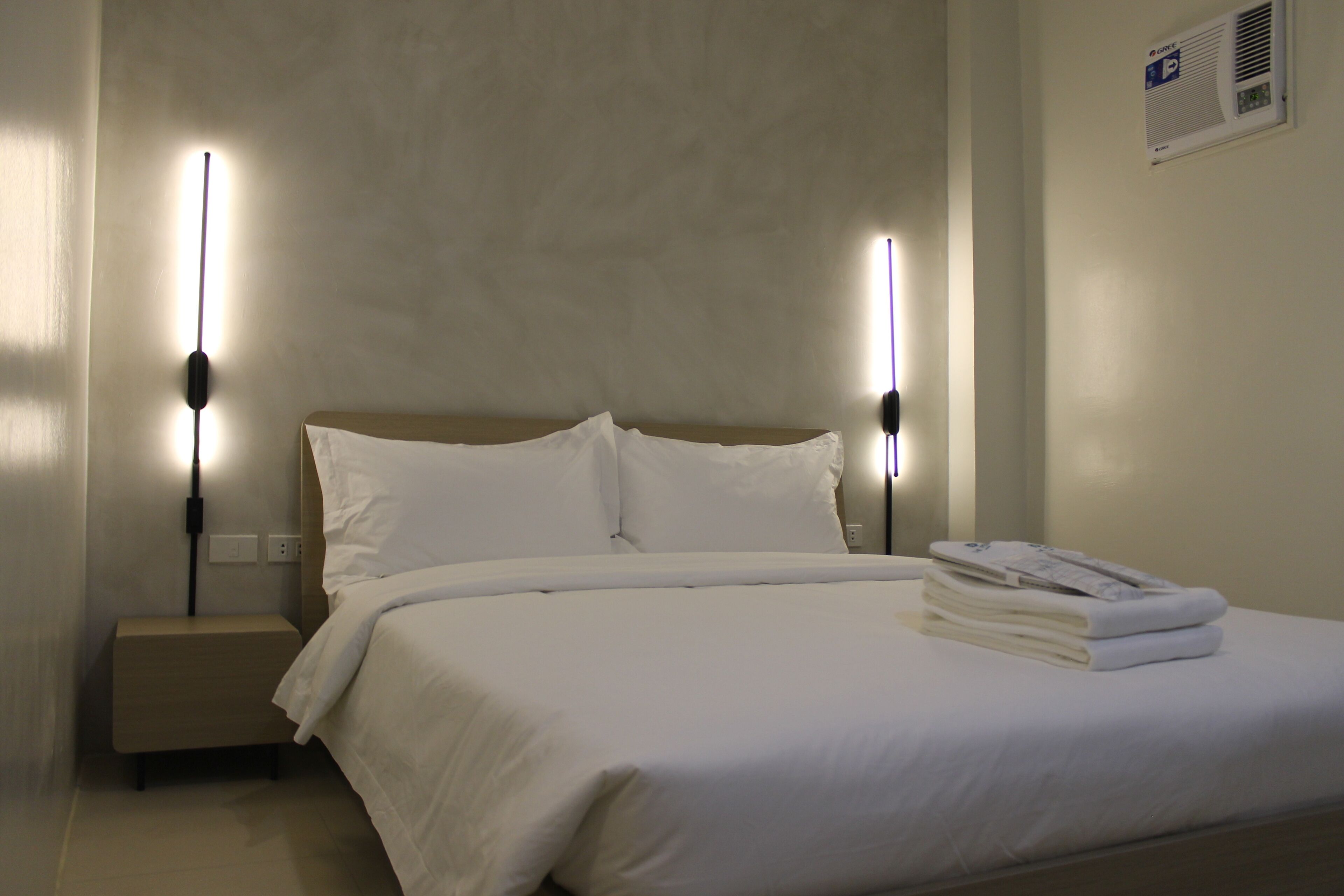 A neatly made bed with white linens, a small bedside table, and two wall-mounted nightstands with lamps.