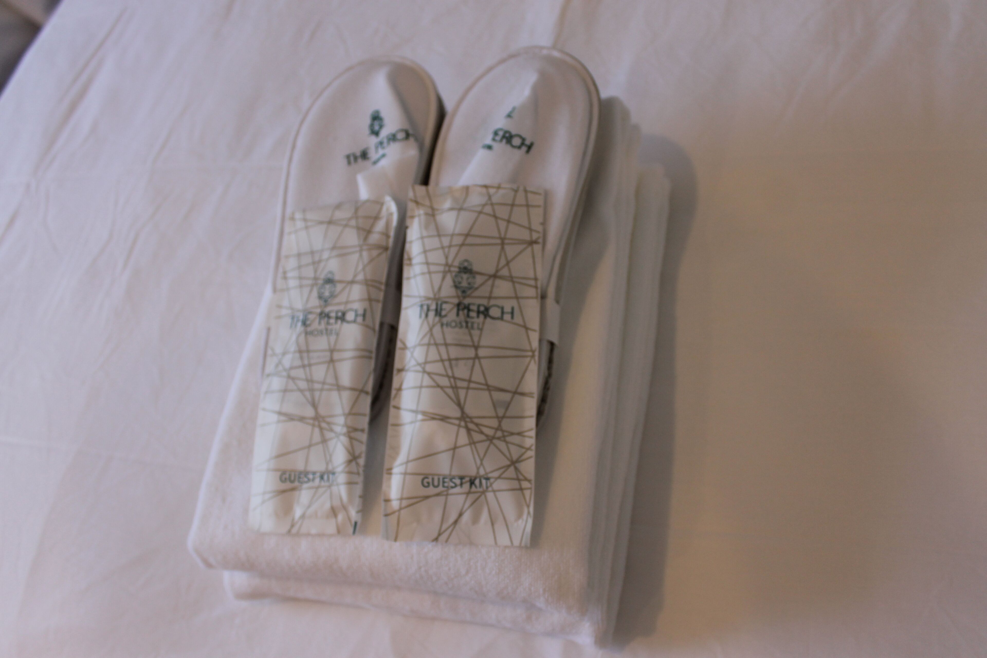 A neatly folded pair of slippers with a geometric pattern and the text "THE MERCH HOSTEL GUEST KIT" on a white bedspread.