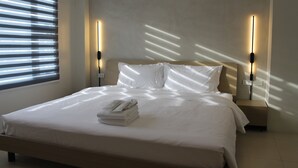 A neatly made bed with white linens and a folded towel, two bedside tables with wall-mounted lights, and a window with blinds. - The Perch Hostel (Olongapo)