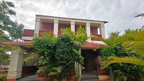 Un edificio de dos pisos con techo de tejas rojas, un balcón con barandillas rojas y un jardín con diversas plantas tropicales.
