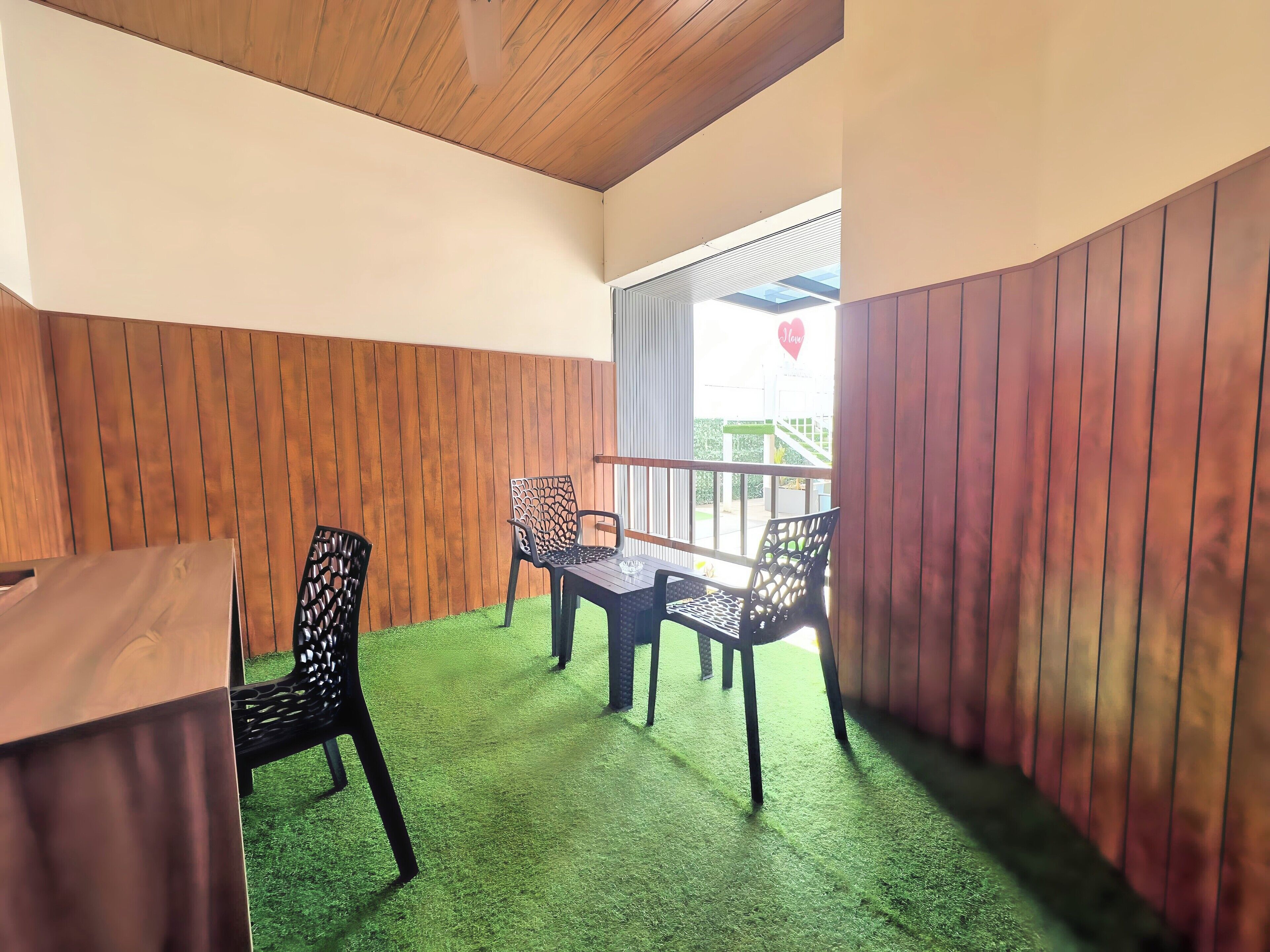 A room with a wooden ceiling, a green artificial grass floor, and a wooden wall. There is a table with chairs and a window with a view outside.