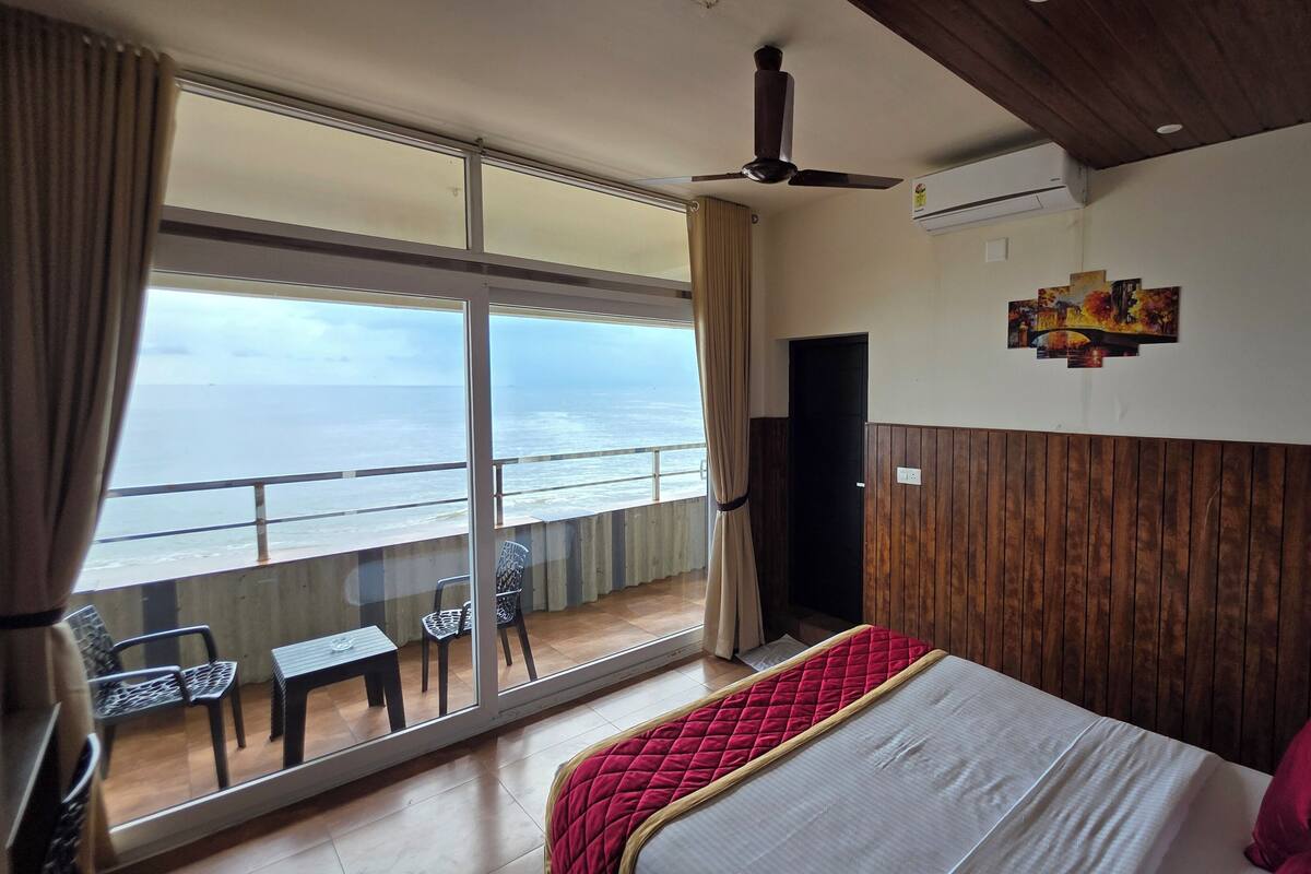 Ein Hotelzimmer mit einer großen Schiebeglasdoor zum Balkon mit Meerblick. Es gibt ein Bett mit roter Steppdecke und weißen Bettwäsche, einen Deckenventilator und eine Wand-Klimaanlage.