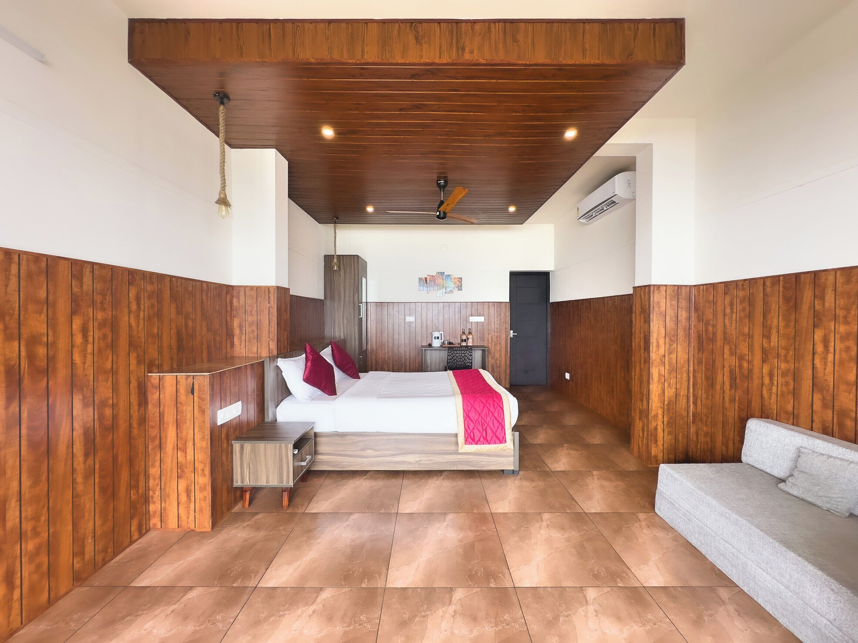 A hotel room with a wooden ceiling, a bed with a red blanket, a grey sofa, and a small wooden nightstand.