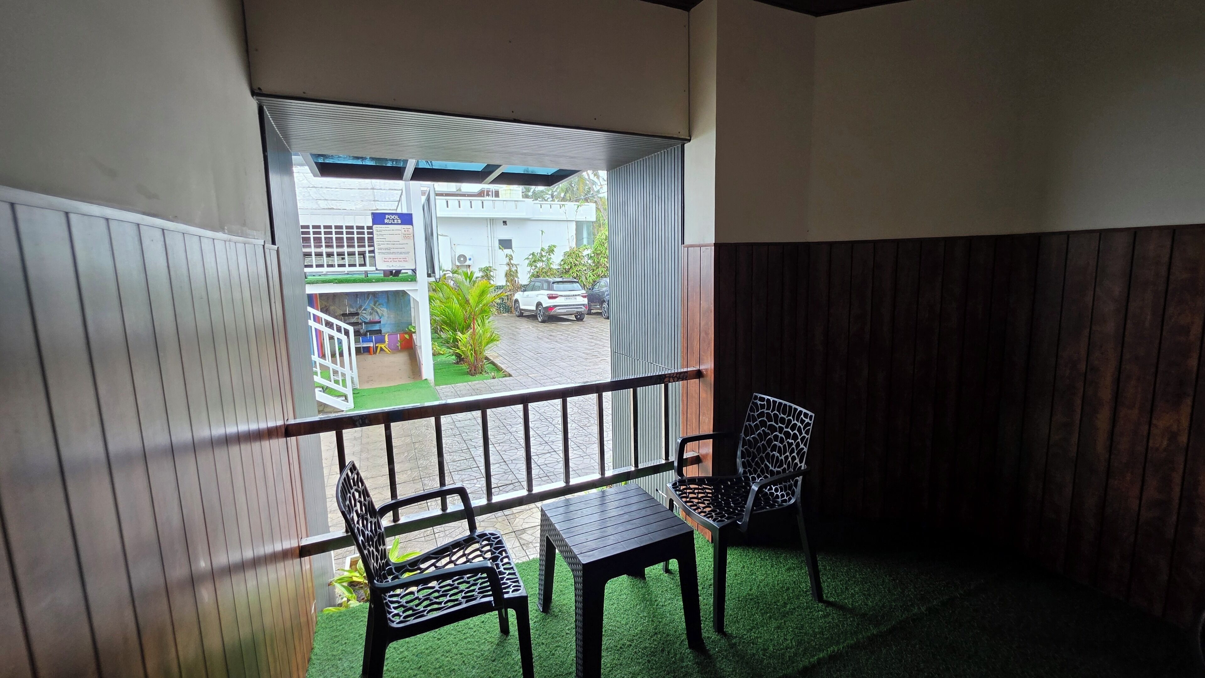 A balcony with two chairs and a table, a view of a street with parked cars, and a building with a sign that reads "POOL RULES".