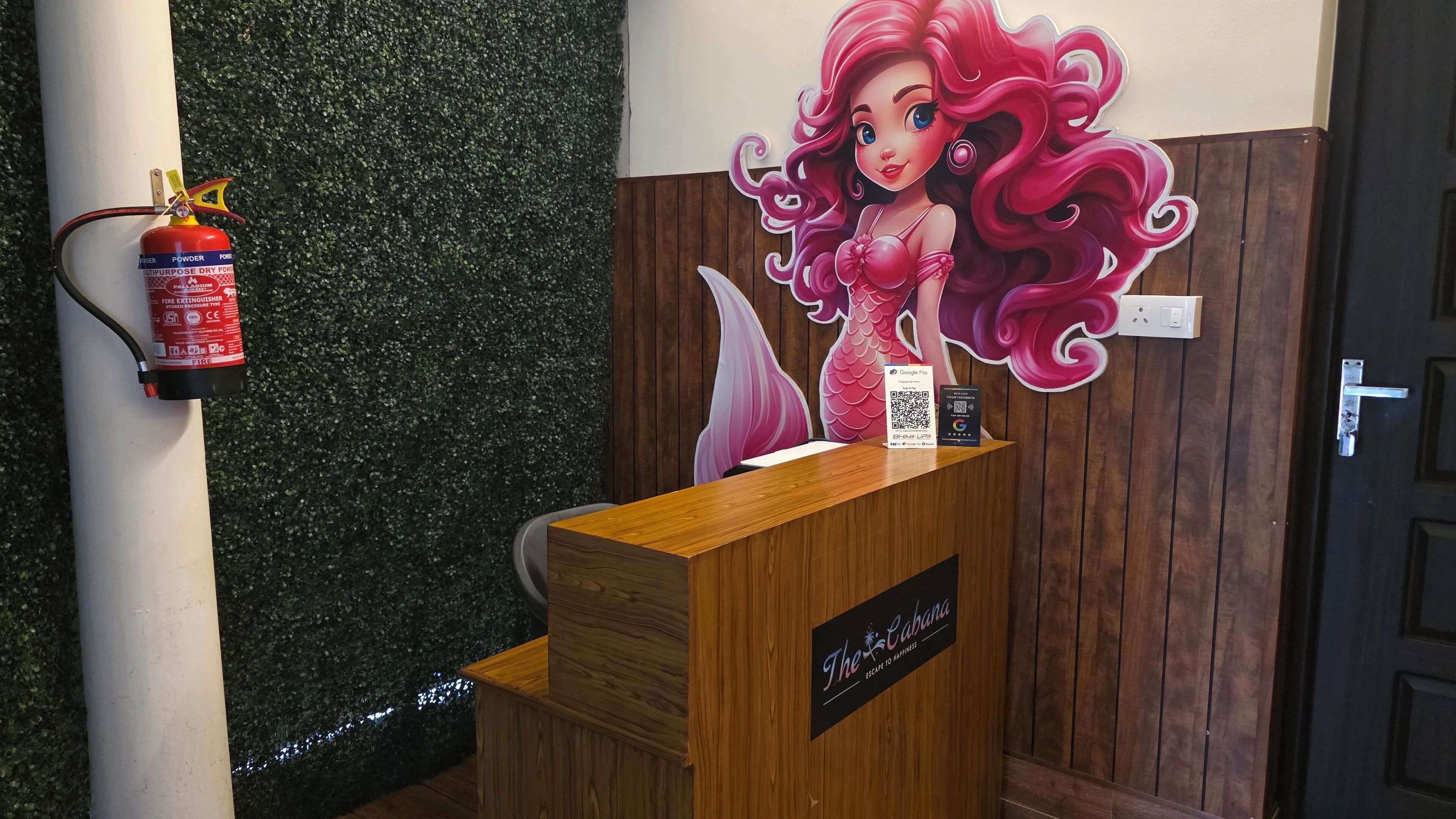 A reception desk with a wooden finish, a fire extinguisher mounted on a white pole, and a mural of a mermaid.