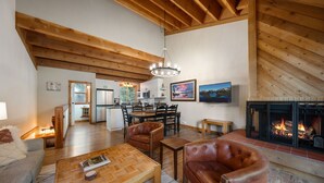 A spacious living area with a fireplace, wooden ceiling beams, a dining table, and a kitchen in the background. - Gold Bend 5008 - Newly Remodeled Dog Friendly Northstar Condo, 2 Suites, HOA Access, Near Trails (Truckee)
