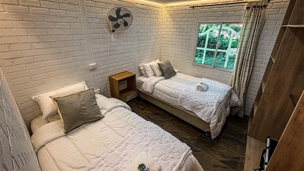A room with two beds, a fan, a window with curtains, and a brick wall. - Betania Cabins (Santa Marta)
